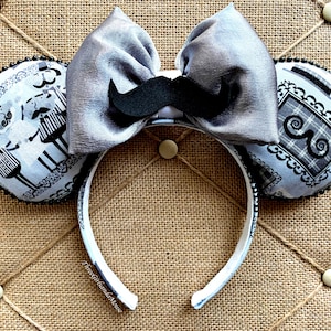 May include: A pair of white mouse ears with a gray satin bow and a black mustache. The ears are covered in a black and white patterned fabric.