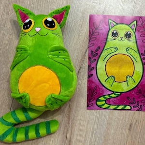 May include: A green plush cat toy shaped like an avocado with a yellow pit. The cat has a striped tail and big eyes. The toy is next to a drawing of a similar avocado cat.