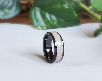 May include: A black ring with a central band of light-colored, speckled material. The ring has a polished, reflective surface and is set against a white background. The ring is a piece of jewelry.