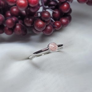 May include: A silver ring featuring a circular, pale pink gemstone. The ring is set against a white backdrop, with a blurred background of red berries. The ring's band is slender and polished.