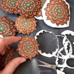 K&ouml;nnte beinhalten: Ein Satz von f&uuml;nf runden Mandala-Aufklebern mit filigranen Designs in Braun-, Orange- und Gr&uuml;nt&ouml;nen. Die Aufkleber befinden sich auf wei&szlig;em Untergrund und wurden mit einem Bastelmesser ausgeschnitten.