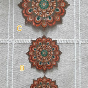 K&ouml;nnte beinhalten: Drei runde Holzmandalas mit komplizierten Mustern, die in Braun-, Orange- und Gr&uuml;nt&ouml;nen bemalt sind. Die Mandalas sind vertikal angeordnet, wobei das gr&ouml;&szlig;te oben und das kleinste unten ist. Die Mandalas sind mit A, B und C beschriftet.