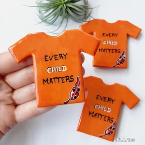 May include: Orange ceramic t-shirt shaped magnets with the text "EVERY CHILD MATTERS" in black and white. Each magnet features a red and black feather design. A small air plant is visible in the background.