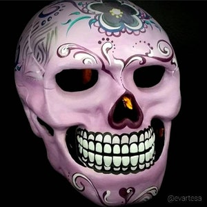 May include: A decorative, light purple skull mask with intricate floral and swirl designs. The mask features large, dark eye sockets and a detailed row of white teeth. The design includes accents of teal, gray, and dark purple, set against a black background.