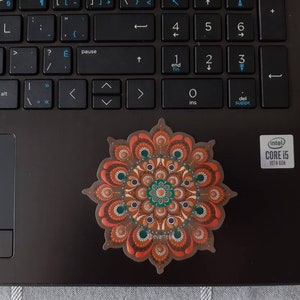 K&ouml;nnte beinhalten: Ein brauner, orangefarbener und gr&uuml;ner Mandala-Aufkleber mit wei&szlig;em Hintergrund. Der Aufkleber befindet sich auf einem schwarzen Laptop.