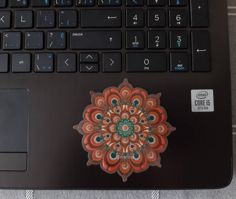 K&ouml;nnte beinhalten: Ein brauner, orangefarbener und gr&uuml;ner Mandala-Aufkleber mit wei&szlig;em Hintergrund. Der Aufkleber befindet sich auf einem schwarzen Laptop.