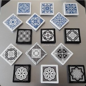May include: A collection of 14 decorative ceramic tiles featuring geometric patterns in shades of black, white, and blue. The tiles are arranged in a grid pattern on a silver surface. All the tiles are square and have a slightly raised edge. The patterns on the tiles are intricate and detailed. These tiles are ideal for adding a touch of style to any room.