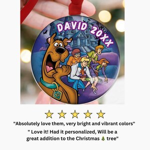 May include: A round Christmas ornament featuring the Scooby-Doo gang in front of a haunted house. The ornament has the name "DAVID" and the year "20XX" printed on it. The ornament is hanging from a red ribbon.