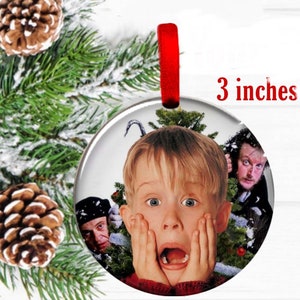 May include: A round Christmas ornament, 3 inches in diameter, featuring a movie scene with a surprised child and two men. The ornament has a red ribbon for hanging and is set against a snowy evergreen backdrop.