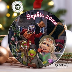 May include: A round, white ceramic ornament featuring a scene from the Muppet Christmas Carol. The characters include Miss Piggy, Kermit the Frog, Gonzo, and Rizzo the Rat. The ornament is personalized with the name "Sophie 20xx".