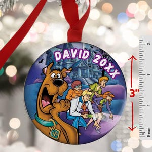 May include: A 3-inch round Christmas ornament featuring the Scooby-Doo gang in front of a haunted house. The ornament has a red ribbon for hanging and includes the text "DAVID 20XX". The ornament is a vibrant, colorful design.
