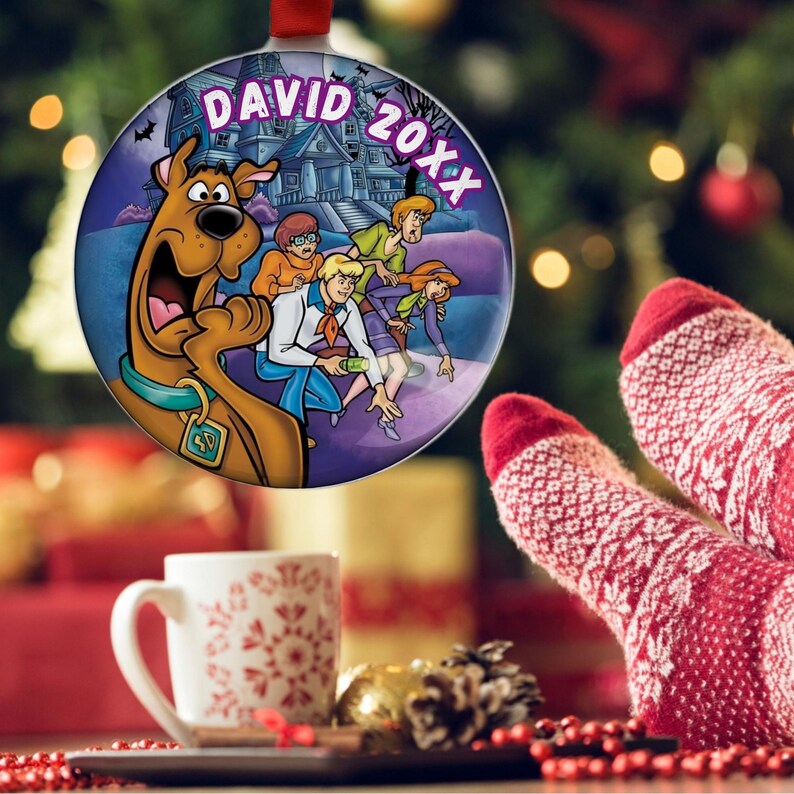 May include: A round Christmas ornament featuring the Scooby-Doo gang in front of a haunted house. The ornament has the text "DAVID 20XX" in white. The ornament is hanging in front of a Christmas tree.