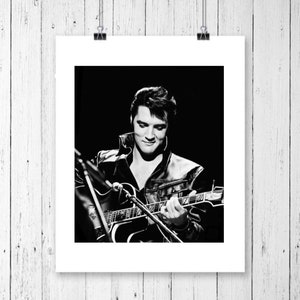 May include: Black and white photo of Elvis Presley playing a guitar. He is wearing a leather jacket and has a microphone in front of him.