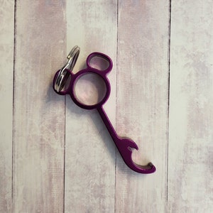 Puede incluir: Abrebotellas de metal morado con un llavero adjunto. El abrebotellas tiene una forma redondeada con un peque&ntilde;o agujero en el centro y un abridor de botellas en la parte inferior.