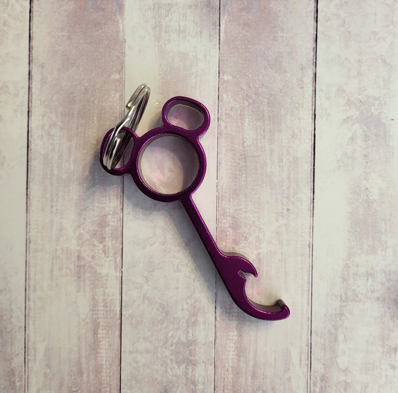 Puede incluir: Abrebotellas de metal morado con un llavero adjunto. El abrebotellas tiene una forma redondeada con un peque&ntilde;o agujero en el centro y un abridor de botellas en la parte inferior.