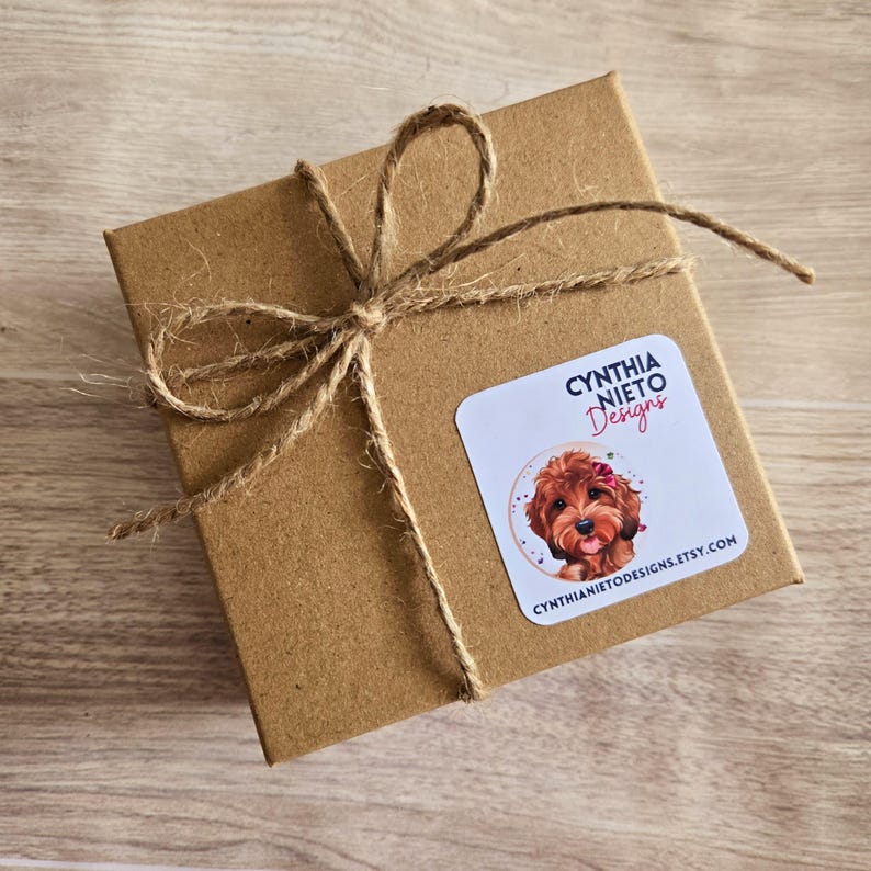May include: A small, square, brown cardboard gift box tied with natural twine. A white sticker on the box features the text "CYNTHIA NIETO Designs" and a watercolor illustration of a dog with a pink bow. The website address is also on the sticker.