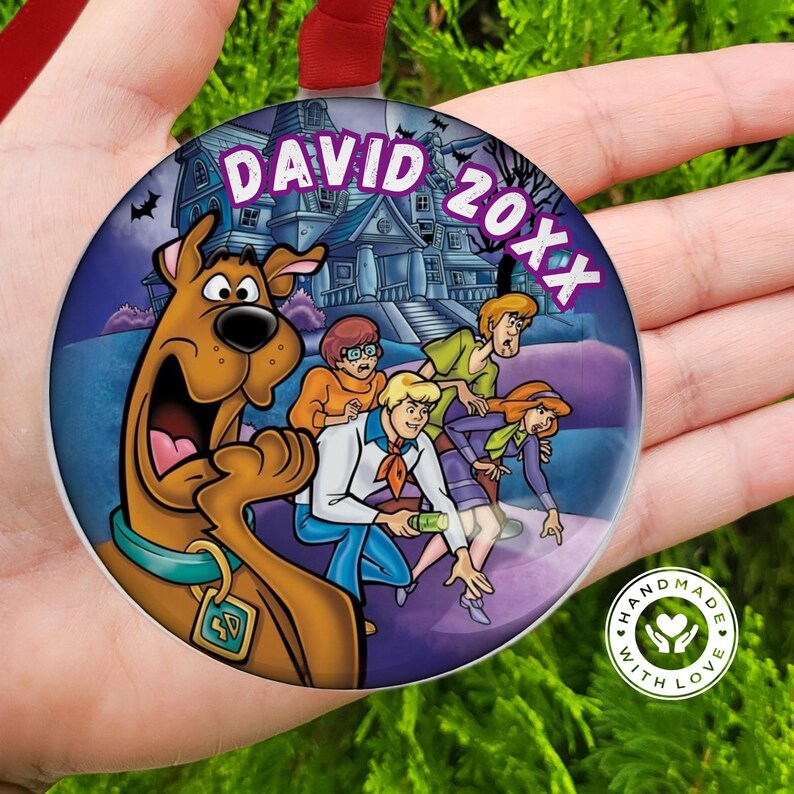 May include: A round Christmas ornament featuring the Scooby-Doo gang in front of a haunted house. The ornament has the name "DAVID" and the year "20XX" in purple text. The ornament is held by a hand.