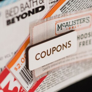 May include: A stack of coupons for Bed Bath & Beyond and McAlister's Deli. The coupons are printed on white paper with black and brown text. The top coupon is for Bed Bath & Beyond and has a red and white $5 off coupon attached to it. The bottom coupon is for McAlister's Deli and has a brown coupon with the word "COUPONS" printed on it.