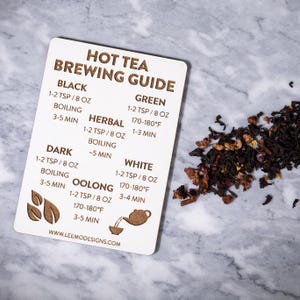 May include: A rectangular wooden tea brewing guide with text and illustrations. The guide provides instructions for brewing black, green, herbal, dark, oolong, and white tea. A pile of loose tea leaves is next to the guide.