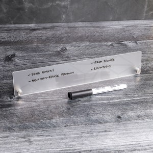 May include: A clear acrylic dry-erase board with a black dry-erase marker. The board has the following text written on it: - Send Emails - New Dry-Erase Product - Prep Dinner - Laundry