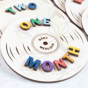 May include: Wooden circle with the words "ONE MONTH" spelled out in colorful letters. The circle has a record-like design with grooves and a smaller circle in the center that says "BABY MEDLOCK".