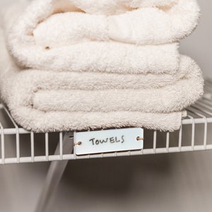 May include: White towels folded and stacked on a white wire shelf with a white label that says "TOWELS" on it.