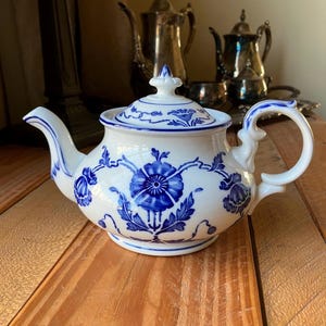 May include: White porcelain teapot with blue floral designs. The teapot features a curved spout and handle, and a lid with a decorative finial. Other silver teapots and serving pieces are in the background. The teapot is sitting on a wooden surface.