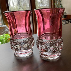 May include: Two clear glass tumblers with a pink rim and interior. The lower portion of the glasses features a decorative, patterned design. The glasses are displayed on a dark brown surface, with a window and plants in the background.