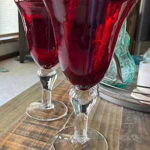 May include: Two ruby red wine glasses with clear stems and bases. The glasses have a flared bowl shape and sit on a wooden surface. The stems have a decorative design. Other glassware is visible in the background.