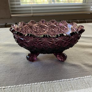 May include: A deep purple glass bowl featuring a pressed starburst and diamond pattern across its exterior surface. The bowl has a scalloped rim and is supported by three small, curved feet, showcasing a translucent finish and geometric design.