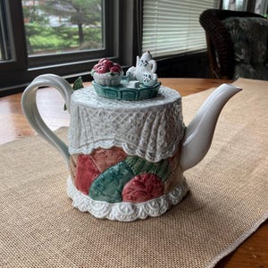 May include: A white ceramic teapot with a decorative lace-like cover and a patchwork design in red, green, and tan. The lid features a miniature tea set and a basket of red berries. The teapot has a curved handle and spout.