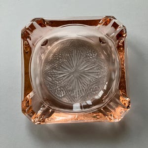 May include: A square, vintage ashtray in a soft pink hue. The ashtray features a central, circular depression with a starburst and floral pattern. The corners are slightly rounded, and the edges are faceted.
