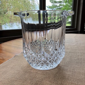 May include: A clear, cut-glass ice bucket with a diamond pattern on the lower half and vertical lines on the upper half. The bucket has two small handles and a round base. A small sticker is visible on the front.