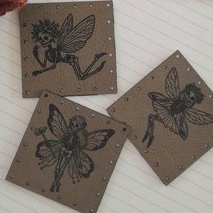 May include: A wooden sign with the text "@WeatheredWeaves" sits on a spiral notebook next to three square leather tags. Each tag features a black fairy illustration. A knitted hat with orange and gray stripes is in the corner.