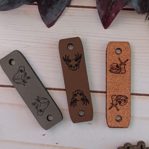 Könnte beinhalten: Drei Holz-Leder-Etiketten mit verschiedenen Tiermotiven. Die Etiketten sind braun und grau und haben oben ein Loch. Der Text "@WeatheredWeaves" befindet sich auf einem Holzschild.
