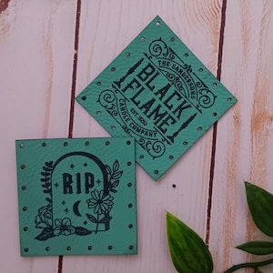 May include: Two teal blue leather patches with black text and designs. One patch says "The Barbershop Black Flame Candle Company Est. 1929" and the other patch says "RIP" with a tombstone and flowers.