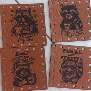 May include: Four square, brown leather patches with raccoon illustrations and text. The patches read: "Jingle Bells & Cozy Vibes", "Sippin' Cocoa & Stealin' Hearts", "Santa? I thought you said Salsa", and "Feral and Festive".