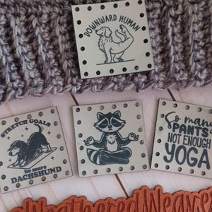 May include: A gray and blue crocheted beanie with four square tags. The tags feature illustrations of a dog, a raccoon, and text that reads "Stretch Goals, be more Dachshund", "Downward Human", and "So many pants not enough yoga".
