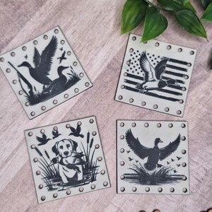 Pode incluir: Quatro bases quadradas, branco-sujo, com ilustrações de patos a preto e branco. Uma base apresenta um pato com um desenho da bandeira americana. As bases são rodeadas por pequenas rebites prateadas. A placa de madeira abaixo diz "@WeatheredWeaves".