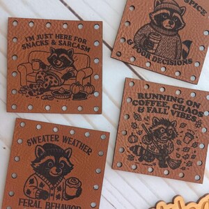 Op de afbeelding: Vier bruine leren patches met wasbeer illustraties en tekst. De tekst luidt: "I'M JUST HERE FOR SNACKS & SARCASM", "PUMPKIN SPICE & POOR DECISIONS", "SWEATER WEATHER FERAL BEHAVIOR" en "RUNNING ON COFFEE, CHAOS & FALL VIBES". Een houten bordje zegt "Weathered Weaves".