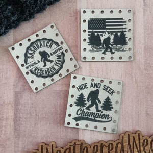 May include: Three metal patches with a Bigfoot theme. The first patch is round and says "Official Sasquatch Research Team". The second patch features a Bigfoot walking in a forest with the American flag in the background. The third patch says "Hide and Seek Champion" with a Bigfoot walking in a forest. The patches are on a wooden surface with a wooden sign that says "@WeatheredWeaves".