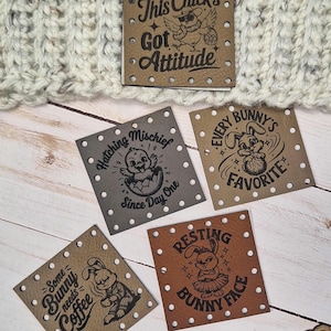 May include: A cream-coloured knitted hat with a brown and white fur pom-pom trim. Several square, brown leather tags with humorous phrases and illustrations are displayed, including "This Chick's Got Attitude."