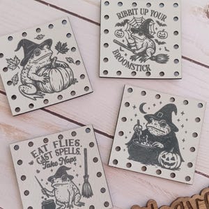 May include: Four square wooden decorations with a Halloween theme. Each features a witch frog illustration with text like "Ribbit up your broomstick" and "Eat flies, cast spells, take naps." A wooden sign with "@WeatheredWeaves" is also present.