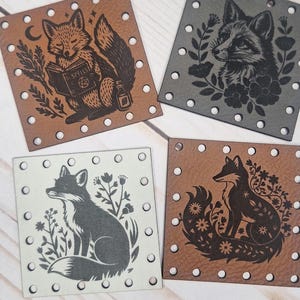 May include: Four square leather patches with fox illustrations. The patches are brown, gray, and off-white, each featuring a different fox design with floral or celestial accents. The patches have small holes around the edges.