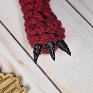 May include: Close-up of a red crocheted dragon foot with three black claws. The foot is made of a textured, dark red yarn. The claws are sharply pointed and appear to be made of a hard, glossy material. The background is a light-colored wooden surface.