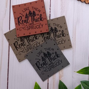 May include: Four square leather tags in shades of brown, olive green, and gray, each with the text "I LIKE THEM Real Thick and Sprucey" in black. The tags have small silver rivets around the edges and are arranged on a white wood surface.