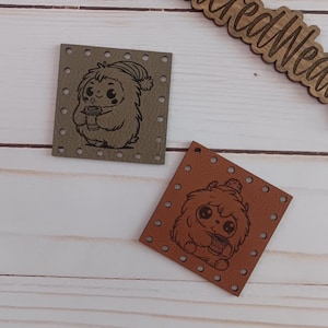 May include: Two small leather patches featuring a cute cartoon creature holding a cup. The creature is wearing a hat and has a fuzzy appearance. The patches are brown and green and have holes around the edges for sewing.