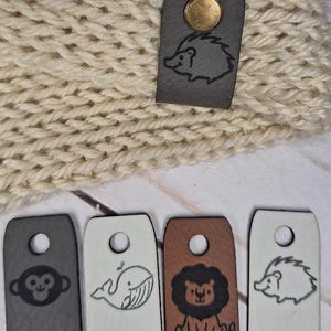 May include: Close-up of a cream-colored knitted hat with a small, dark gray leather tag featuring a gold rivet and a hedgehog illustration. Below are several leather tags with animal illustrations, including a monkey, whale, lion, and hedgehog.