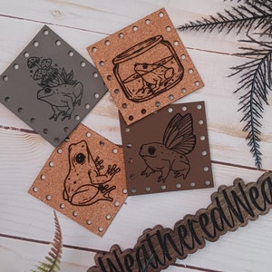 May include: Four leather patches featuring a frog theme. The patches are brown and depict a frog in a jar, a frog with wings, a frog sitting on a leaf, and a frog with a mushroom. The patches are decorated with small holes around the edges. The patches are labelled "@WeatheredWeaves".