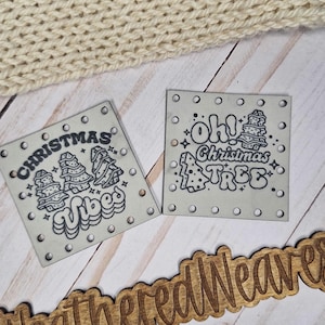 May include: Two small, square, off-white Christmas-themed signs with black line art and text, and a wooden sign with the text "@WeatheredWeaves". A cream-colored knitted item is in the background.